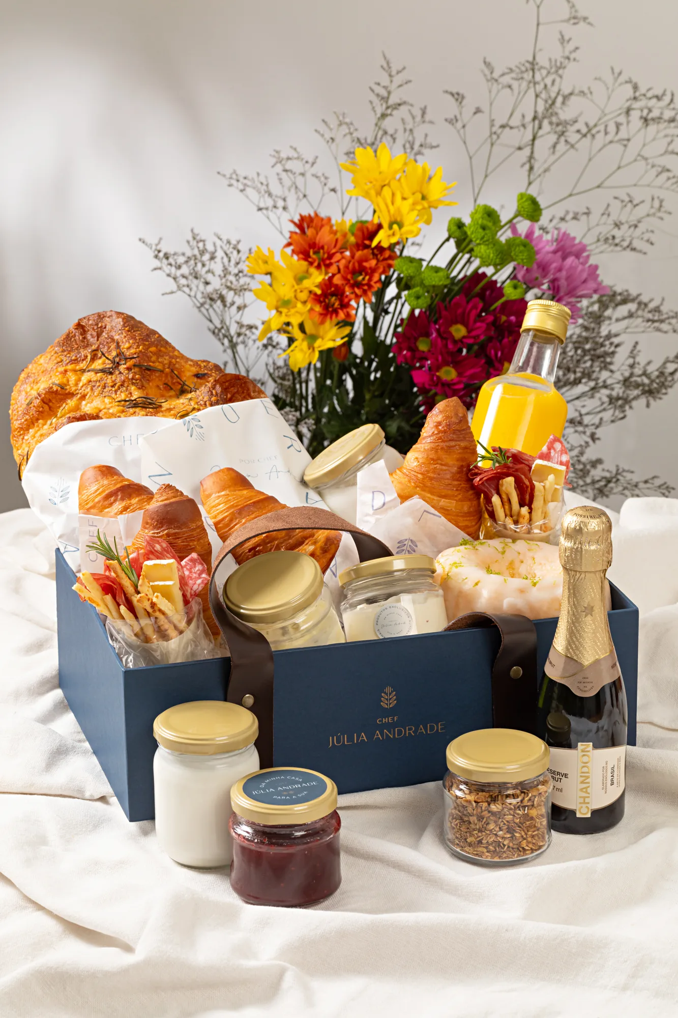 Cesta brunch montada na caixa azul da marca, com focaccia, croissants, sucos e flores