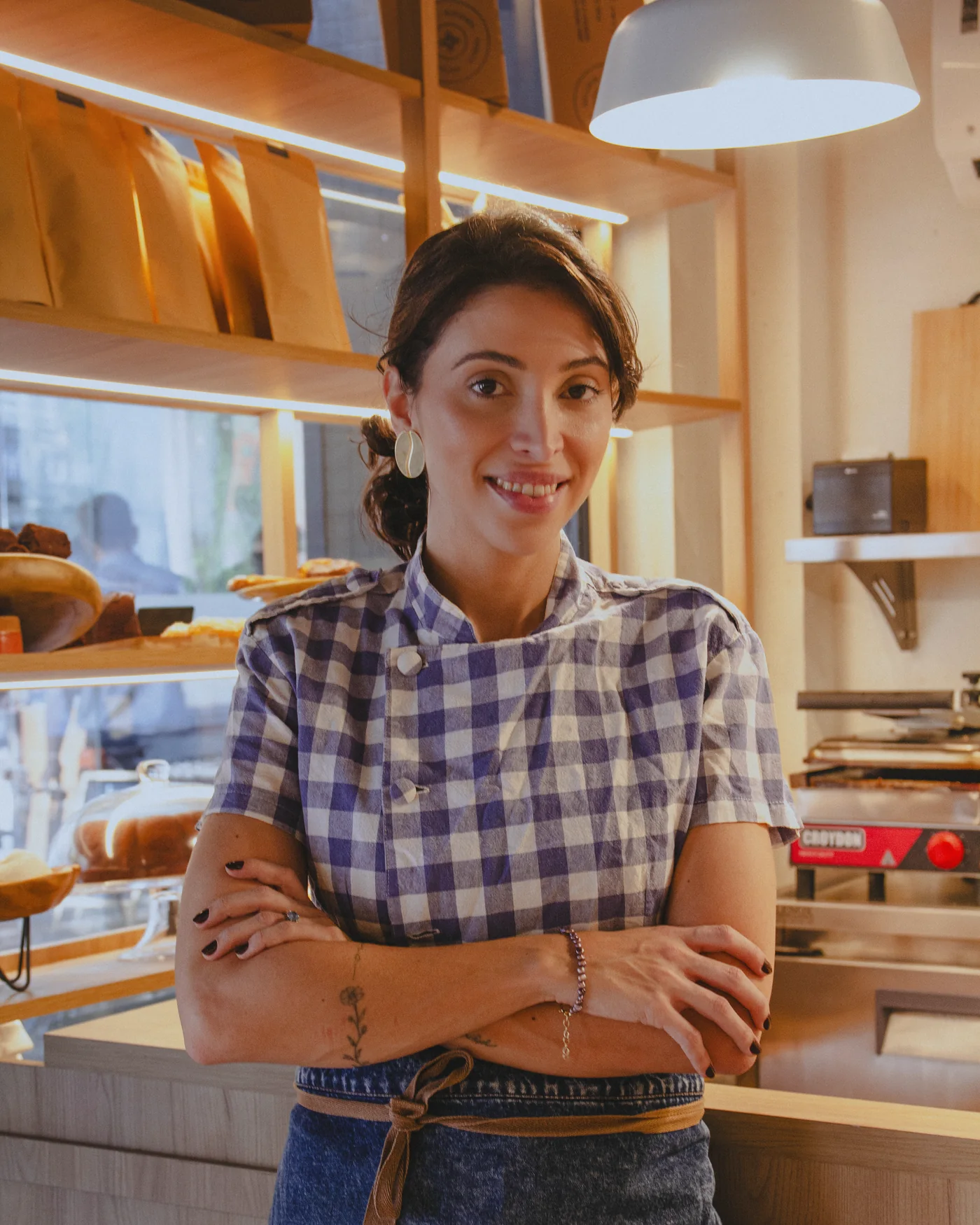 Júlia Andrade de braços cruzados atrás do balcão da DÉLI, de camisa xadrez azul e avental jeans, com a vitrine de pães e as embalagens da casa na prateleira