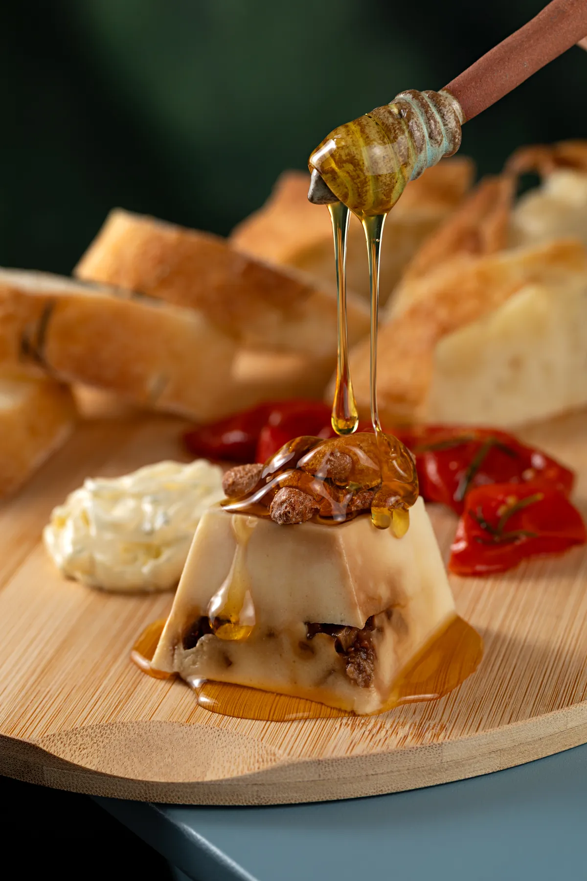 Mel escorrendo de um pegador de madeira sobre uma mousse com castanhas caramelizadas, numa tábua com fatias de focaccia, creme e pimentões assados