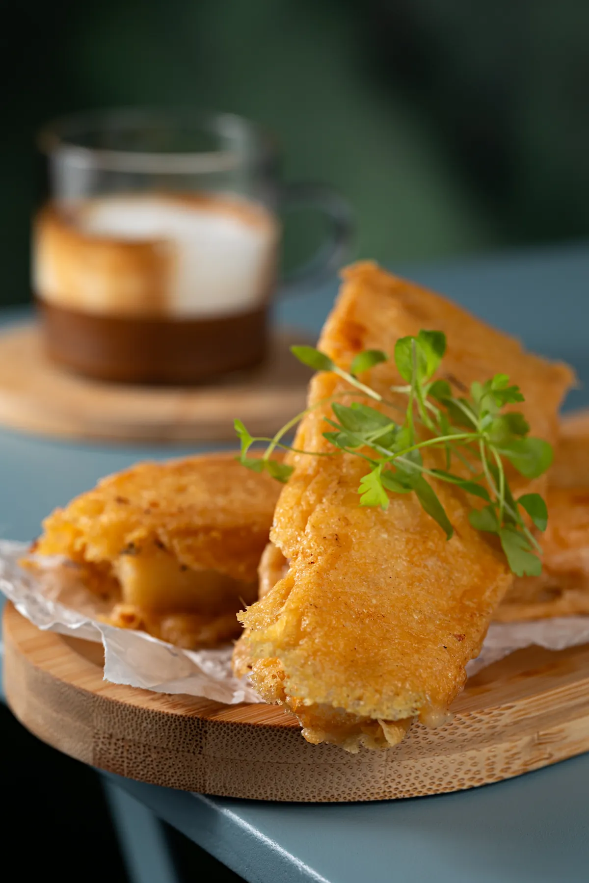 Fatias de focaccia tostadas e douradas, com crosta de queijo e brotos por cima, ao lado de um café com leite em copo de vidro