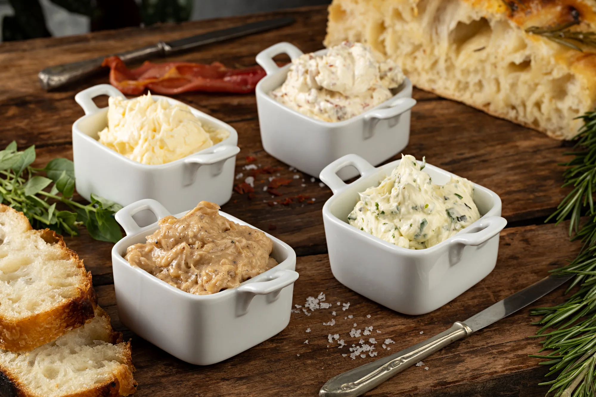 Quatro tigelinhas brancas com manteigas e pastas diferentes sobre tábua de madeira, ao lado de fatias de pão, focaccia e ervas frescas