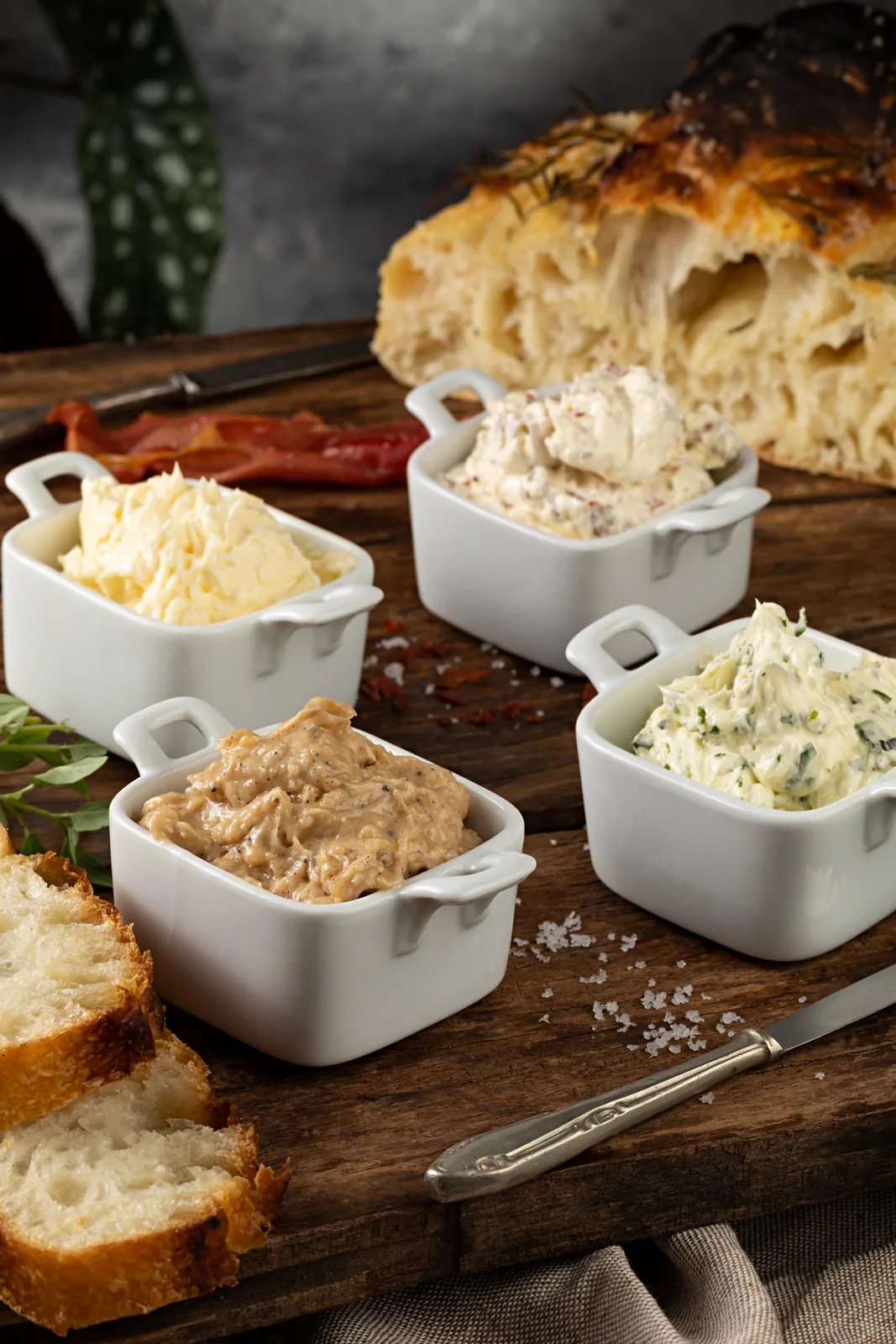 Quatro potes brancos com pastas e manteigas diferentes, ao lado de focaccia e pão fatiado sobre tábua de madeira