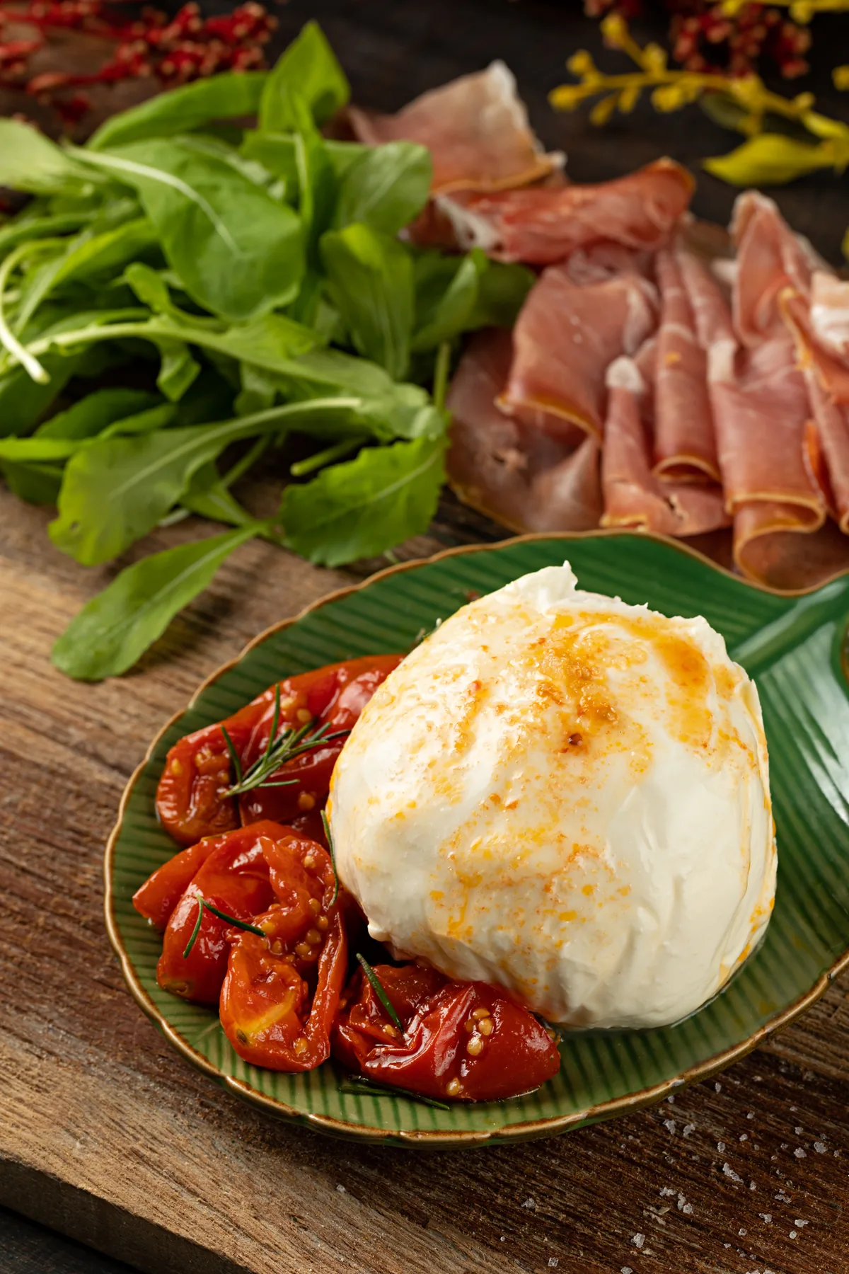 Burrata inteira sobre folha verde, com tomates assados, presunto de parma e rúcula ao lado